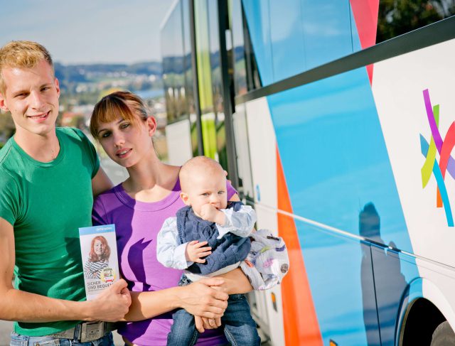 Familie mit Baby hält Öffi-Ticket vor einem Bus im Salzburger Land. Nachhaltig reisen.