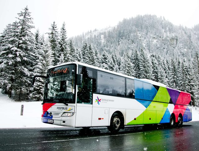 Bunter Postbus vor verschneiten Bäumen im Salzburger Land, ideal für nachhaltige Reisen.